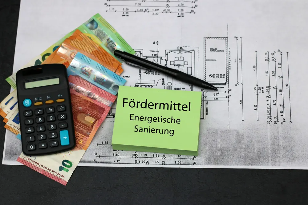 Taschenrechner, Euroscheine und Bauplan mit Zettel „Fördermittel Energetische Sanierung“